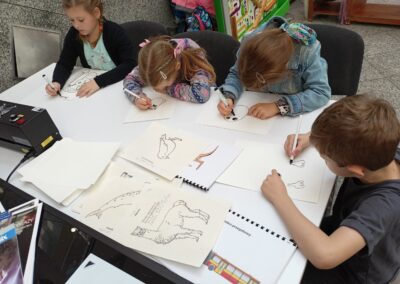 Czworo dzieci przy stole rysuje obrazki na papierze do wygrzewarki. Na stole znajduje się również wygrzewarka, która spowoduje, że obrazki staną się wypukłe.