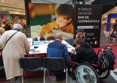 Stoisko WOMAI na akcji Sensorycznie i na serio: stół przy którym siedzą wolontariusze i uczestnicy warsztatów, na pierwszym planie chłopiec na wózku