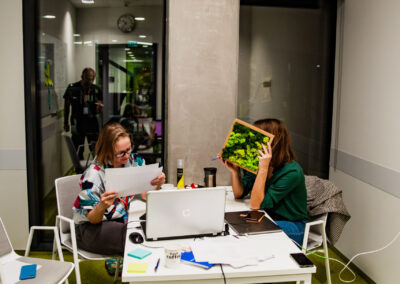 Sala w biurowcu, przy stole siedzą dwie kobiety, uczestniczki hackathonu, jedna z nich zasłania twarz zieloną tabliczką. Pracują nad projektem.