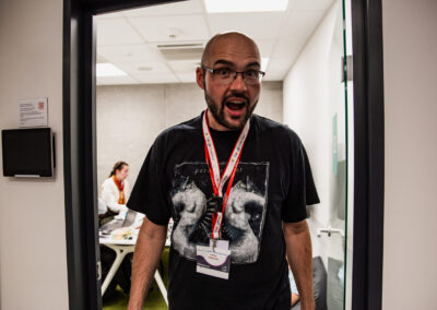 Mężczyzna w koszulce metalowej wychodzi z pokoju, w którym pracują uczestnicy hackathonu i robi zabawną minę do kamery, która mówi "o nie :)"