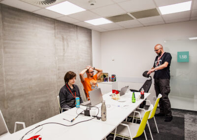 Sala w biurowcu, przy stole siedzi dwoje uczestników hackathonu, obok stoi mentor. Pracują nad projektem.