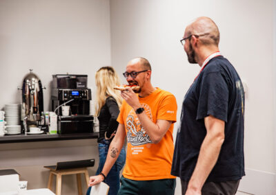 Kuchnia w biurowcu, na pierwszym planie dwóch uczestników hackathonu, jeden z nich zjada kawałek pizzy, na drugim planie ekspres do kawy i kobieta, która czeka na swoją kawę.