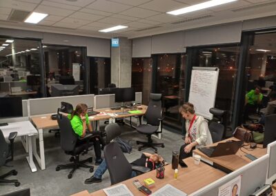 Trzy osoby w dużej pustej sali biurowej, dwie siedzą na krzesłach biurowych, jedna leży na podłodze. Pracują nad projektem. Jest noc.