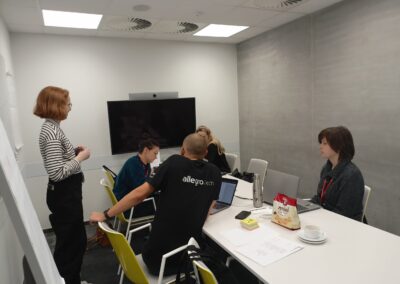 Sala w biurowcu, przy stole siedzi czworo uczestników hackathonu, obok stoi kobieta. Pracują nad projektem.