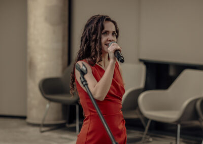 Kobieta z długimi, kręconymi włosami, elegancko ubrana w czerwoną sukienkę z mikrofonem w ręku. Śpiewaczka podczas występu.