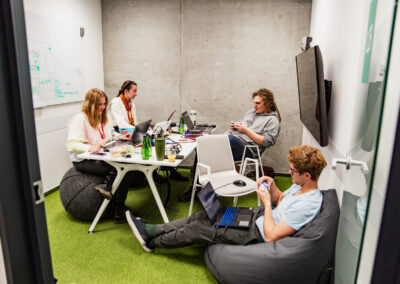 Sala w biurowcu, przy stole siedzi troje uczestników hackathonu, obok na puffie siedzi czwarty mężczyzna. Pracują nad projektem.