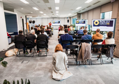 Sala w biurowcu na której odbywa się konferencja widziana od tyłu - ludzie siedzą na krzesłach, mężczyzna i kobieta stoi przy ekranie multimedialnym