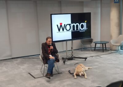 Mężczyzna siedzi na krześle przy ekranie multimedialnym z napisem WOMAI, obok przy jego nogach leży biszkoptowy pies przewodnik